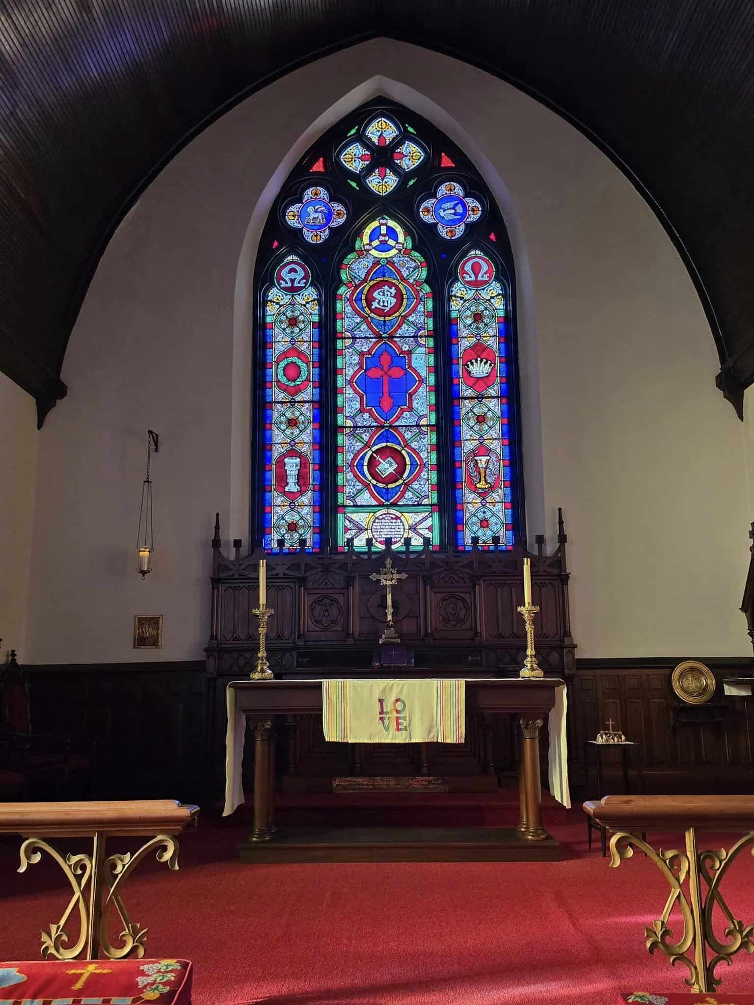 Church interior with ornate stained glass windows and wooden altar. Red carpet leads to altar with candles and cloth inscribed with "LOVE".