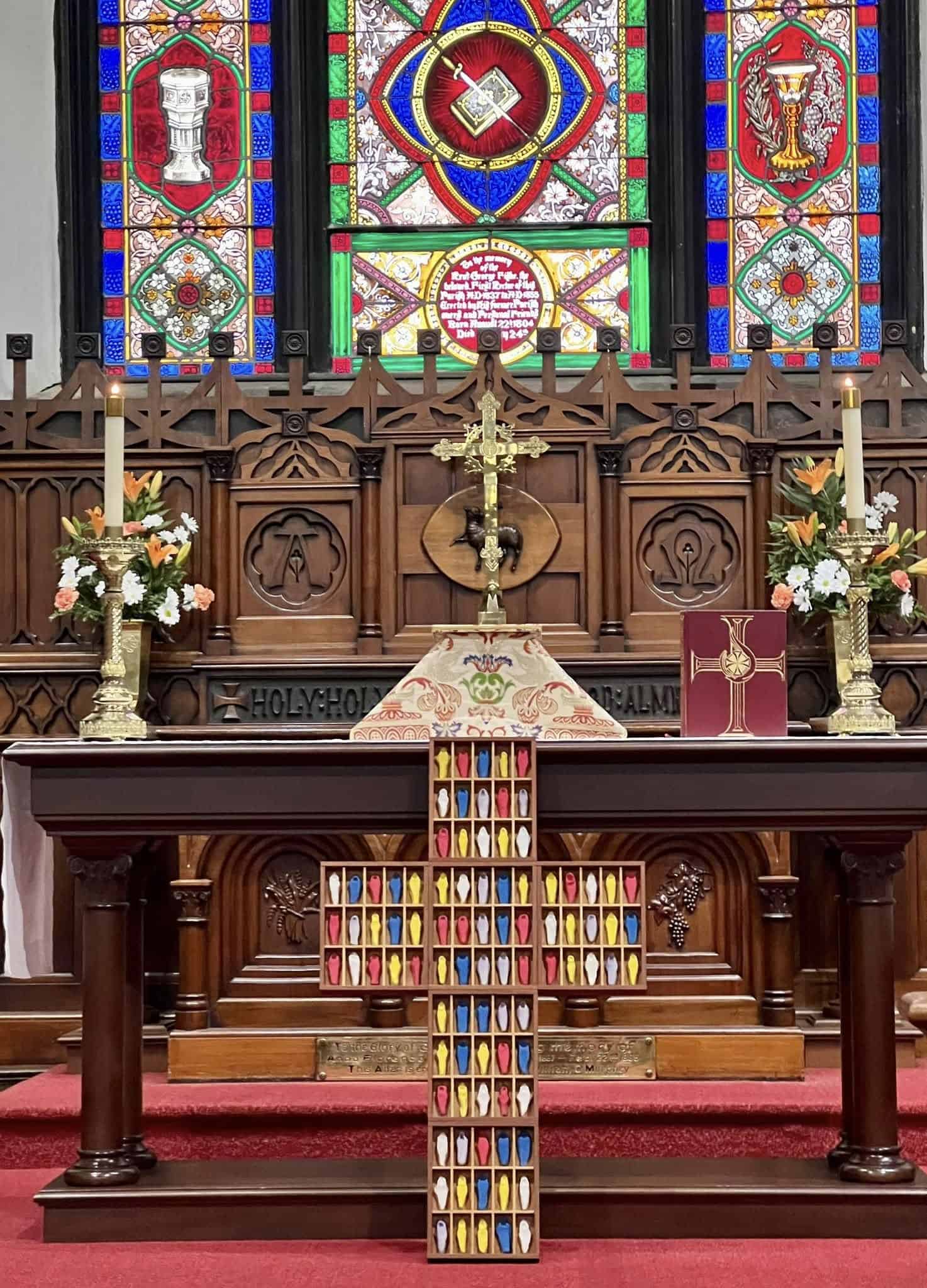 Ornate church altar with colorful stained glass, wooden cross with candles, floral arrangements, and religious symbols. Richly decorated space suggests a solemn atmosphere.