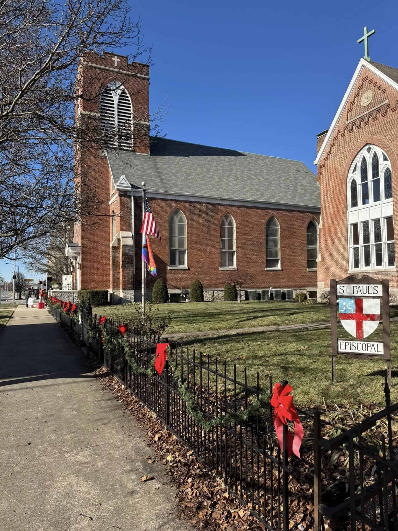 St. Paul's Episcopal Church, a brick building with arched windows, is adorned with festive decorations and flags, next to a sidewalk.